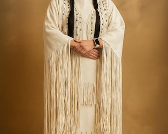 May include: A woman in a cream-coloured Native American dress with fringe and beadwork. She has long black plaits and a feather in her hair. The dress has long sleeves and a matching skirt. She is wearing a smartwatch.