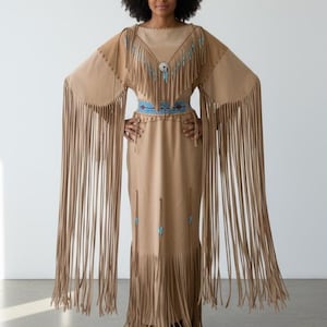 May include: A tan suede dress with long fringe sleeves and hem. The dress features a beaded neckline and belt with turquoise accents. The sleeves are long and flowing, and the dress has a simple, elegant silhouette.