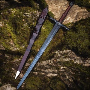 Puede incluir: Una espada de estilo medieval con una larga hoja plateada y un mango marrón. La espada tiene una guarda negra y está acompañada por una vaina de cuero marrón. Los objetos se encuentran sobre un fondo verde musgoso.