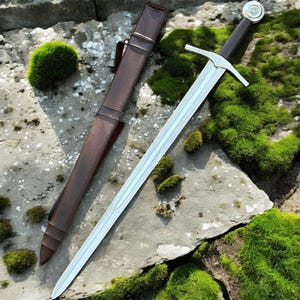May include: A medieval-style sword with a silver blade and a brown handle, resting on a stone surface with green moss. The sword is accompanied by a brown leather sheath. The hilt features a circular emblem.