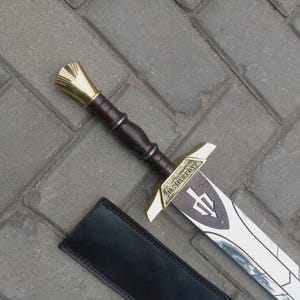 Op de afbeelding: Een decoratief zwaard met een goudkleurige pareerstang en kruisbeschermer, een bruin handvat en een wit lemmet met een drietandsymbool. Het zwaard ligt op een grijs bakstenen oppervlak naast een zwarte schede.