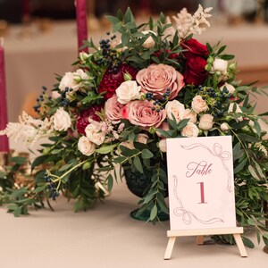 May include: A floral centerpiece with red, pink, and white roses, greenery, and berries. A table number card with the word "Table" and the number "1" is displayed. Burgundy candles and gold candle holders are also present.