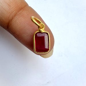 May include: A small, rectangular pendant featuring a deep red gemstone set within a gold-coloured metal frame. The gemstone's surface is faceted, reflecting light. The pendant has a small loop at the top for attaching to a chain or bracelet.