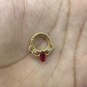 Puede incluir: Un anillo de tabique dorado con una gema roja oscura en forma de lágrima en el centro. El anillo presenta delicados detalles de filigrana y pequeñas piedras de acento claras.
