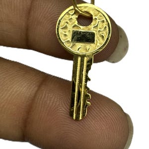 May include: A gold-colored key pendant with a textured circular head and a rectangular detail. The key's stem has intricate grooves and a loop for a chain. The pendant is held between fingers against a white background.