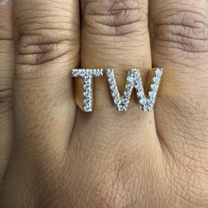 Puede incluir: Anillo dorado con las letras "TW", cada una incrustada con brillantes gemas blancas. El anillo es una pieza audaz, que muestra un diseño moderno con un toque de lujo. Las gemas están muy juntas, creando una exhibición brillante.