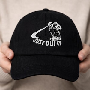 May include: Black baseball cap with a white graphic of a golfer swinging a club. The text "JUST DUI IT" is printed in white below the graphic. The cap is made of a soft material and has a curved brim.
