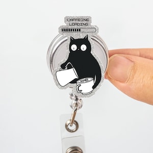 Puede incluir: Un carrete retráctil para placa con un gato negro vertiendo café en una taza. El diseño incluye el texto "CAFFEINE LOADING" encima de una barra de progreso. El carrete tiene un borde plateado brillante y un clip de plástico transparente.