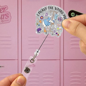 Op de afbeelding: Een glitter badge reel met de tekst "I PICKED THE WRONG POLE". De badge heeft een cartoon eend in een medische setting. De reel heeft een doorzichtige plastic band en een metalen clip.