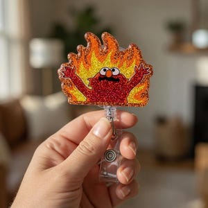 May include: A red glitter badge reel featuring a cartoon character engulfed in flames. The character has large eyes, a black mustache, and outstretched arms. The badge reel is held by a clear plastic clip.
