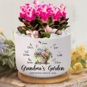 May include: A white ceramic planter with a vibrant pink flowering plant. The planter is decorated with a floral design, butterflies, and the text "Grandma's Garden GROWING SINCE 2026". It sits on a wooden coaster.