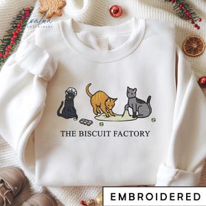 May include: White sweatshirt with an embroidered design featuring three cartoon cats. The cats are in various poses, with the text "THE BISCUIT FACTORY" below. The word "EMBROIDERED" is at the bottom.