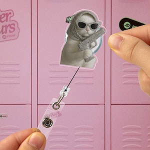 Puede incluir: Un gato blanco con gafas de sol y un pañuelo con pedrería, sosteniendo una pistola cubierta de pedrería. La imagen del gato está en un portatarjetas retráctil con una correa de plástico transparente y un clip de metal. El fondo es un casillero rosa.