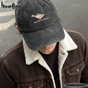 Puede incluir: Gorra de béisbol gris oscuro con un diseño bordado de un ganso rosa con una rana verde en la espalda. El texto "we ride at dawn" está debajo del ganso. La gorra la lleva una persona con una chaqueta marrón.