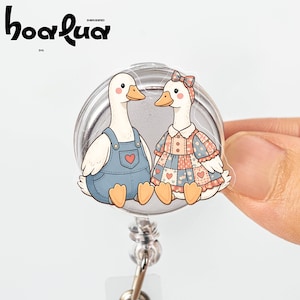 May include: A badge reel featuring two cartoon ducks. One duck wears blue overalls with a heart, the other a dress with a bow. The badge reel has a silver metal backing and a retractable cord. The logo "hoalua" is at the top.
