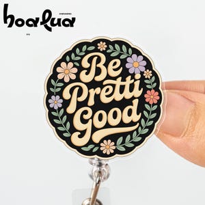 May include: A round badge reel with the words "Be Pretti Good" in a cream-colored, retro font. The badge is black with floral accents in peach, lavender, and green. The badge reel has a clear plastic cover and a silver-colored metal clip.