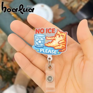 May include: A badge reel with a cartoon cat illustration. The badge reel has the words "NO ICE PLEASE" and a drawing of a glass of ice water. The cat is orange and white with a grumpy expression.