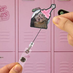 Puede incluir: Un carrete retráctil para placa con un mono sonriente que lleva un gorro de fiesta rosa. El carrete tiene una correa de plástico transparente y un clip metálico. El fondo es un casillero rosa.