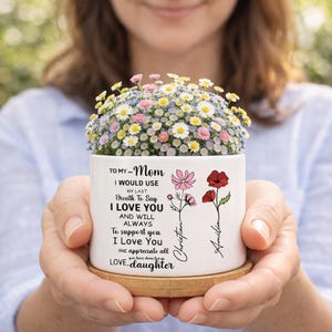 May include: A white ceramic planter filled with colorful flowers, held in a person's hands. The planter has a message for "Mom" with the names "Christian" and "Amilia" and a bamboo coaster.