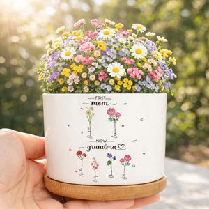 May include: A white ceramic flower pot with the words "FIRST mom - NOW grandma" and floral illustrations. The pot is filled with a vibrant mix of colorful flowers, including daisies and other blooms. The pot sits on a round wooden base.