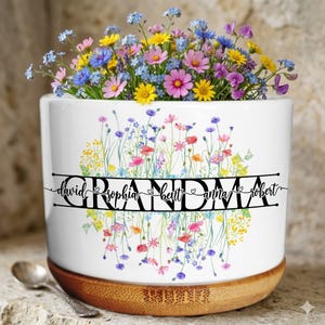 May include: A white ceramic planter with a floral design and the word "Grandma" in black script. The planter is filled with colorful flowers and sits on a wooden base. The names David, Sophia, Beitt, Anna, and Robert are also included.