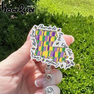 May include: A Mardi Gras-themed badge reel featuring a map of Louisiana in purple, green, and yellow. The badge reel has text that says "Fleur-de-lis", "Mardi Gras", "Beads", "King Cake", and "Crawfish".