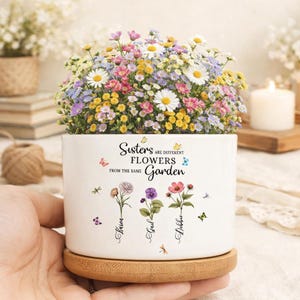 May include: A white ceramic planter with a wooden base, filled with colorful flowers. The planter has the text "Sisters are different flowers from the same garden" and three names. A hand is holding the planter.