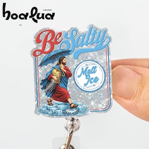 May include: A badge reel with a religious illustration. The image features a figure holding a bag of salt under an umbrella, with the words "Be Salty" and "Melt Ice". The design has a silver glitter background and a blue and red color scheme.