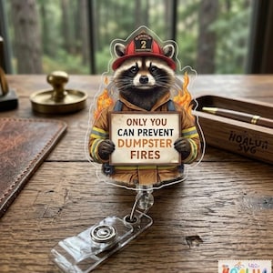Könnte beinhalten: Ein klarer Acryl-Ausweis-Rollenhalter mit einem Waschbären, der als Feuerwehrmann verkleidet ist und einen roten Helm mit der Nummer 2 trägt. Der Waschbär hält ein Schild mit der Aufschrift "ONLY YOU CAN PREVENT DUMPSTER FIRES". Das Design umfasst Flammen und eine transparente Kunststoffrolle.