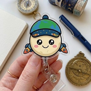 Puede incluir: Un portatarjetas de acrílico transparente con un personaje de luna de dibujos animados que lleva un gorro verde y azul con el tema de la Tierra. La luna tiene una cara sonriente, mejillas rosas y detalles de cohetes. El portatarjetas tiene un cordón retráctil y un clip metálico.