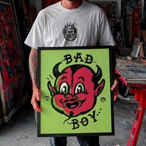 May include: A framed artwork featuring a cartoon devil face with the words "BAD BOY" in black. The devil's face is red with black horns and a green background. The artwork is held by a person wearing a white t-shirt.
