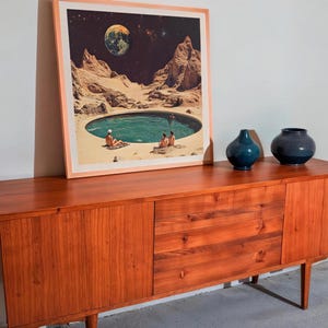 a wooden cabinet with a framed picture of a desert landscape with a pool of water in the center. The cabinet has two vases on top of it.