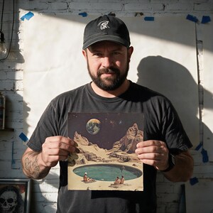 A man with a beard is holding up a framed picture of a surreal landscape with a pool and mountains, against a white brick wall.
