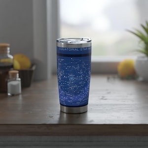 Op de afbeelding: Een roestvrijstalen reismok met een blauwe achtergrond met een wit sterrenkaartontwerp. De tekst "EQUATORIAL STAR MAP" staat bovenaan. De mok is cilindrisch en heeft een zilveren deksel.