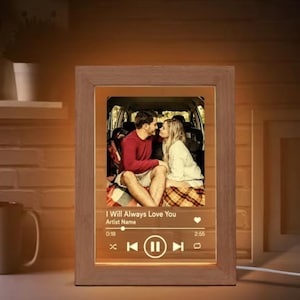May include: A wooden framed photo with a couple kissing inside a car. The photo is illuminated with a warm glow. Below the photo is a music player interface with the text "I Will Always Love You".