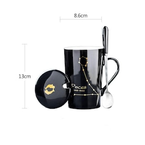 May include: Black ceramic mug with a lid and spoon. The mug is 13cm tall and 8.6cm wide, featuring a gold constellation design and the word "Pisces". The lid has a gold fish symbol. A silver spoon rests on the side.