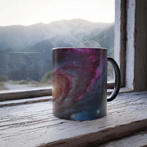 Puede incluir: Una taza de café con asa negra que presenta un diseño de galaxia colorido con tonos morados, azules y naranjas. Sale vapor de la taza, lo que sugiere una bebida caliente. La taza está sobre un alféizar de madera blanca desgastada.