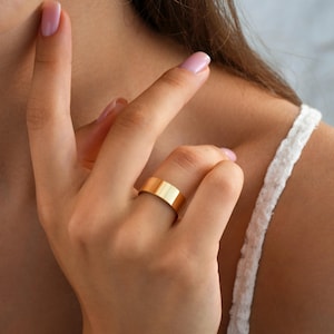 May include: A gold-colored ring with a flat, rectangular design. The ring is worn on a finger. The ring is a simple, elegant piece of jewelry.