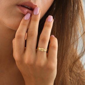 May include: A gold ring featuring a woven design. The ring is on a finger, with a hand and part of a face in view. The ring's detailed pattern is the main focus, displaying a mix of sophistication and artistry. The ring is a beautiful piece of jewellery.