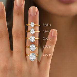 May include: Five gold rings with oval-cut clear stones in varying sizes, labeled 1.00 ct, 1.50 ct, 2.00 ct, 3.00 ct, and 5.00 ct. Each ring features a thin, polished gold band and a prong setting holding a single oval-shaped gemstone.