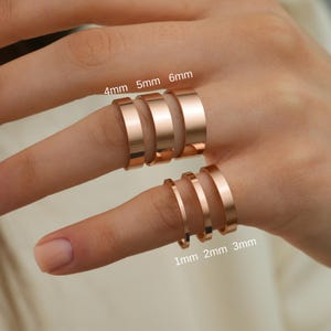 May include: Six rose gold-toned rings with flat, smooth surfaces, displayed in varying widths. The rings are labeled with text indicating widths of 1mm, 2mm, 3mm, 4mm, 5mm, and 6mm, showing the comparative thickness of each band on the fingers.
