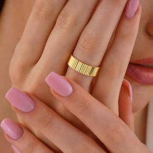 May include: A gold ring with a ribbed design. The ring is worn on a finger. The ring's surface has vertical lines, creating a textured appearance. The ring is a simple band style.