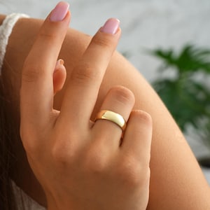 May include: A gold-colored ring with a smooth, rounded design. The ring is worn on a finger, showcasing its polished surface and simple elegance. The ring's warm hue contrasts with the skin tone, highlighting its classic appeal.