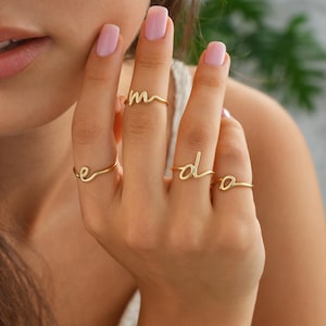 Peut inclure: Quatre bagues dorées délicates, chacune conçue avec une lettre cursive : 'e', 'm', 'd' et 'o'. Les bagues sont présentées sur une main aux ongles peints, mettant en valeur la simplicité élégante des bijoux. Les bagues sont un accessoire élégant.