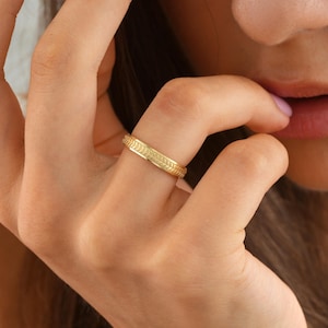 May include: A gold ring with a detailed leaf pattern. The ring is worn on a finger, showcasing its intricate design and polished finish. The ring's band features a repeating pattern, adding to its elegance.