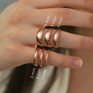 May include: A selection of rose gold rings in various widths, from 1mm to 6mm, are shown on fingers. The rings have a polished finish, with sizes clearly labelled. The image showcases the different ring sizes available.