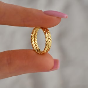 May include: A gold ring featuring a repeating leaf pattern. The ring is held between two fingers, highlighting its detailed design and polished surface. The ring's design is a continuous pattern of stylized leaves.