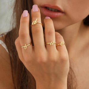 Peut inclure: Quatre bagues dorées, chacune ornée d'une lettre cursive différente : 'd', 't', 'a' et 'b'. Les bagues sont présentées sur une main aux ongles vernis. Les bagues sont délicates et semblent être en métal brillant.