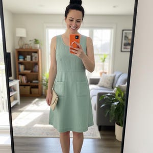 May include: A sleeveless, light green dress with two front pockets. The dress has a scoop neckline and a knee-length hem. A small, beige clutch is held in one hand. The dress is made of a textured fabric.