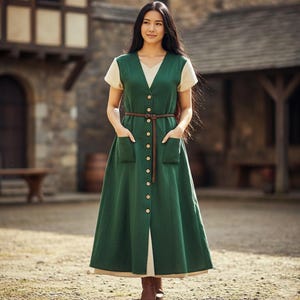 May include: A green button-down dress with a V-neck and short sleeves. The dress features a brown belt and two front pockets. The dress is layered over a cream-colored top and is paired with brown boots. The dress is a long, flowing style.